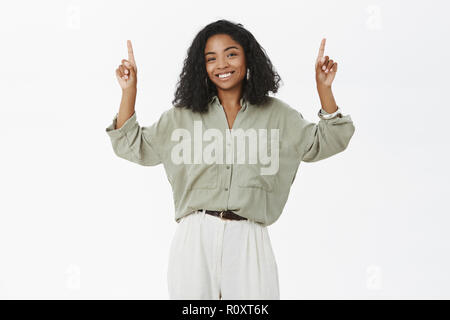 Affascinante di carnagione scura modello femminile con capelli ricci nel quartiere alla moda di camicia e pantaloni i bracci di sollevamento e rivolta verso l'alto sorridente cordiale e felice di consigliare lo spazio di copia per i clienti Foto Stock