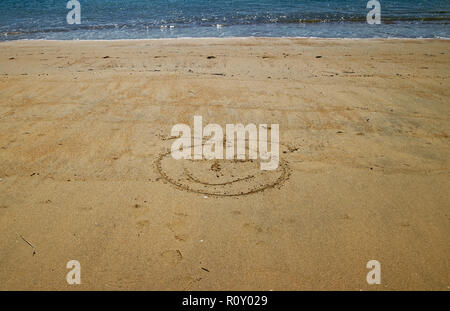 Un disegno che ti faranno sorridere, una faccina sorridente nella sabbia dorata con mare blu dietro Foto Stock