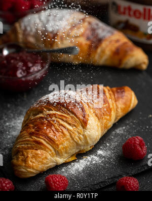 Croissant essendo spolverato con zucchero a velo circondato con lamponi Confettura di lamponi e nutella Foto Stock