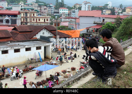 Due giovani ragazzi crouch su una parete per guardare oltre la vendita di piccoli animali a Bac Ha mercato domenicale, Vietnam Foto Stock