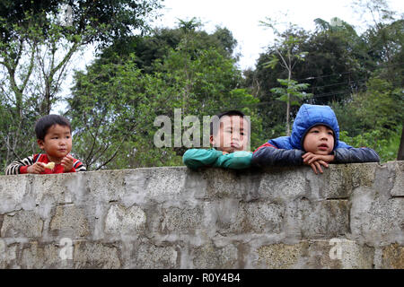 Tre giovani ragazzi peer su un blocco in calcestruzzo a parete, Ban Pho village, Vietnam Foto Stock