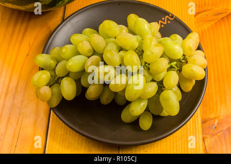 Still Life - un pennello grande di uva verde e anguria in un buio piastra ceramica su uno sfondo di legno Foto Stock