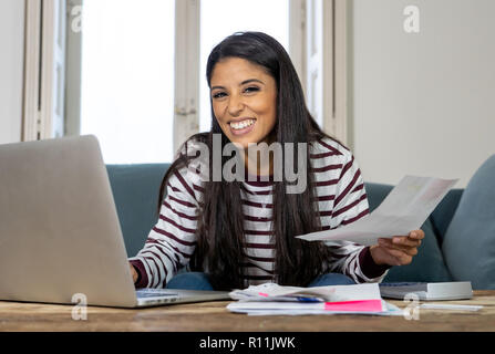 Attraente sorridente donna latina pagare le bollette costi contabili imposte tasse e mutui ipotecari a casa utilizzando la calcolatrice portatile e cercando contento e soddisfatto Foto Stock