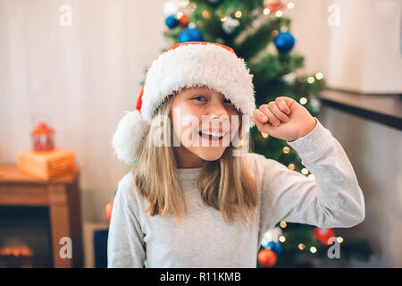 Allegro e positivo ragazza sorridente e ridere. Essa detiene il bordo del suo cappello di Natale. La ragazza si erge nella stanza dove si decora albero di Natale Foto Stock