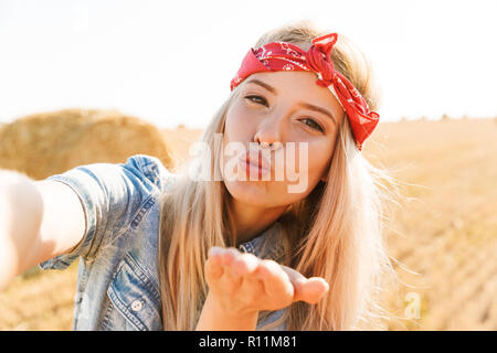 Bella giovane sorridente ragazza bionda in fascia al campo di grano, prendendo un selfie, invio di kiss Foto Stock