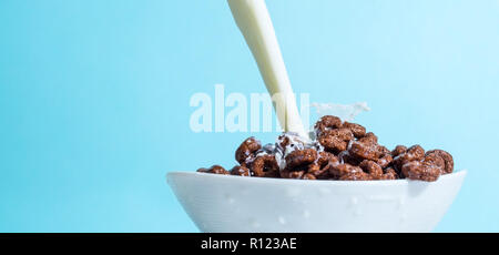 Flusso di latte versando in una tazza con scaglie di cioccolato in forma di anelli, spruzzi di latte su sfondo azzurro. Foto Stock