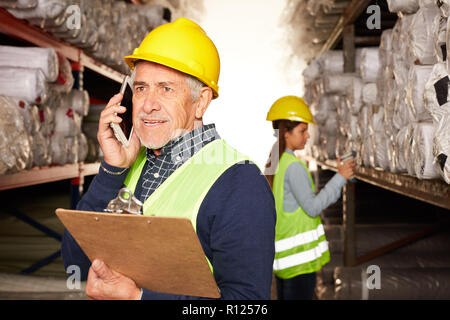 Vecchio lavoratore logistica telefona con lo smartphone nel magazzino di tappeti Foto Stock