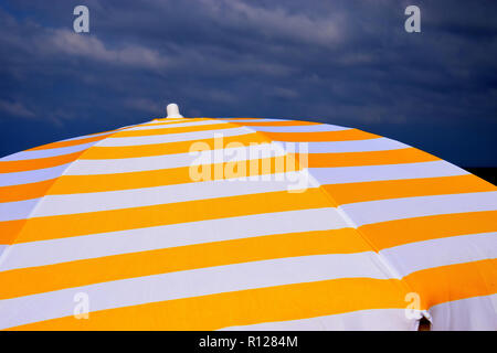 Cielo nuvoloso e ombrellone in spiaggia, giallo e bianco striato nella visiera parasole anteriore del cielo nuvoloso come segno di cattivo tempo di vacanza Foto Stock