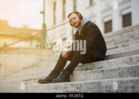 Un bel giovane hipster uomo seduto sulle scale e guardando la telecamera. Foto Stock