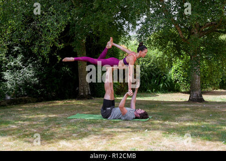 Giovane uomo a praticare yoga in giardino, giacente sul retro donna di bilanciamento su piedi Foto Stock