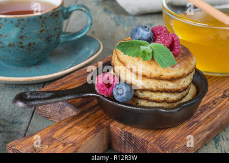 La colazione in stile rustico: frittelle in ghisa padelle, miele e frutti di bosco freschi e il tè nero Foto Stock