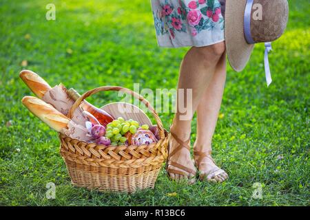 Estate - picnic sul prato nel parco. ragazza e cesto per un picnic con baguette, vino, bicchieri, uva e rotoli Foto Stock