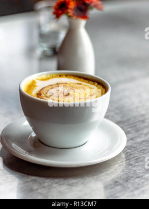 Casual caffetteria. Latte art. La formazione di schiuma del latte. Brocca in vetro chiaro. Fiori d'arancio in un bianco ovale vaso bianco. Foto Stock