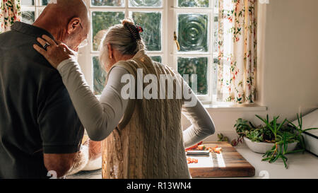 Vista posteriore di una coppia senior in piedi insieme nella cucina di cottura degli alimenti. Donna anziana guardando suo marito cucinare il cibo in piedi in cucina. Foto Stock