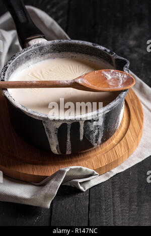Latte bollito in una pentola nera e rovesciato sopra i bordi, un cucchiaio di legno su un tagliere, su un tavolo rustico. Luce bassa dell'immagine. Foto Stock