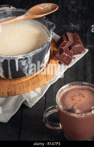 Il latte bollito in una pentola nera, rovesciato sopra i bordi, hunks di cioccolato e una tazza di vetro con cioccolata calda, su un tavolo rustico. Foto Stock