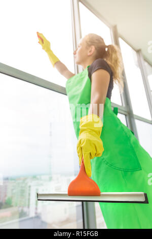 Sorridente donna giovane lavoratore la pulizia schiuma di sapone sulla finestra in vetro con spatola e rag Foto Stock