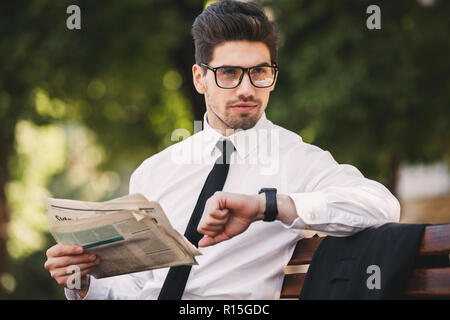 Foto di grave imprenditore nel soddisfare la lettura di giornale sul banco in parco verde e guardando un orologio da polso Foto Stock