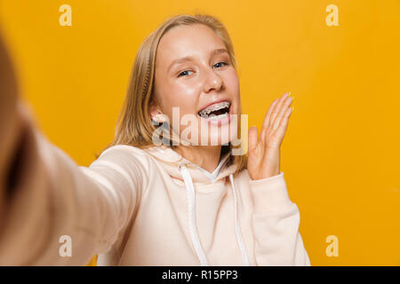 Foto di allegra ragazza 16-18 con bretelle dentali sorridente e tenendo selfie isolate su sfondo giallo Foto Stock