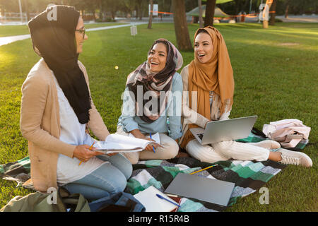 Foto di un felice giovani donne arabe degli studenti che utilizzano computer portatili e contenere libri nel parco. Foto Stock