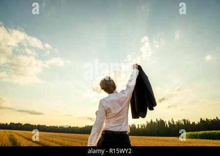 Vista da dietro di un imprenditore in piedi nel campo segato al di sotto del cielo della sera con il suo braccio di sostegno suit giacca sollevato in alto al trionfo o celebrazione. Foto Stock
