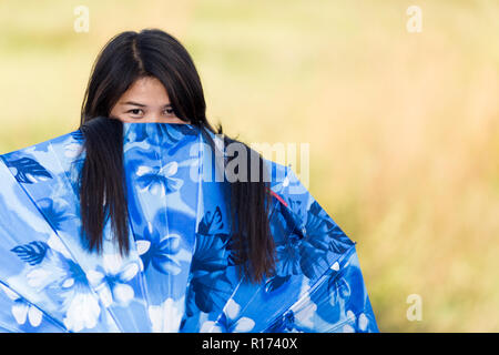 Giocoso giovane ragazza tailandese peeking sopra la parte superiore del suo blue Ombrello o ombrellone con un divertito sguardo come lei gioca fuori nel sole caldo, con copia Foto Stock