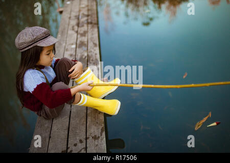 Bambino ragazza in giallo stivali di gomma e cappuccio si siede sul legno ponte di pesca e le catture di pesce con self-made canna da pesca. Vista dall'alto. Foto Stock