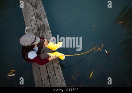 Bambino ragazza in giallo stivali di gomma e cappuccio si siede sul legno ponte di pesca e le catture di pesce con self-made canna da pesca. Vista dall'alto. Foto Stock