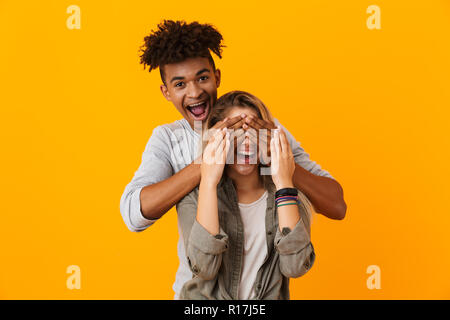 Ritratto di una felice coppia multietnica in piedi isolato su sfondo giallo, coprendo gli occhi della sua fidanzata Foto Stock