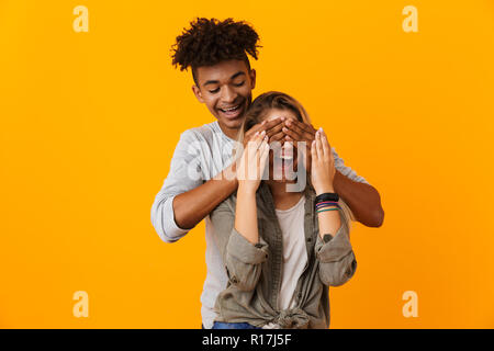 Ritratto di un sorridente coppia multietnica in piedi isolato su sfondo giallo, coprendo gli occhi della sua fidanzata Foto Stock