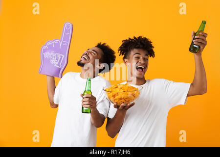 Due gioiosa uomo africano amici isolate su sfondo giallo guardando la partita di calcio, a bere birra e mangiare patatine Foto Stock