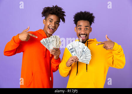 Due maschi felici amici africani in colorate felpe con cappuccio in piedi isolato su sfondo viola, mostrando il denaro banconote Foto Stock