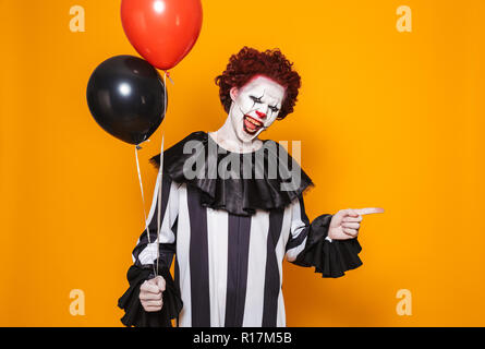 Angry Man vestito in scary clown costume di Halloween palloncini di contenimento isolato su sfondo giallo Foto Stock