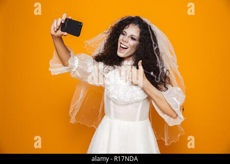 Immagine di gioiosa sposa zombie di halloween indossando abiti da sposa e trucco spaventoso tenendo selfie sul telefono cellulare isolato su sfondo giallo Foto Stock