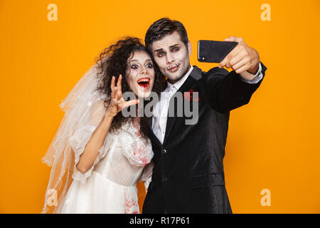 Foto di gioiosa zombie giovane sposo e sposa che indossa il vestito di nozze e Halloween trucco ridere mentre prendendo selfie isolate su backg giallo Foto Stock