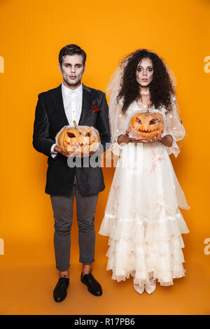 A tutta lunghezza shot di giovani creepy halloween sposa e lo sposo in costumi di nozze guardando la fotocamera e tenendo la zucca in web isolate su Orange Foto Stock