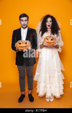 A tutta lunghezza shot di sorpreso scary halloween sposa e lo sposo in costumi di nozze guardando la fotocamera e tenendo la zucca in web isolate su Orange Foto Stock