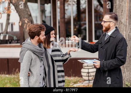 Amichevole maschio agente immobiliare di consegnare la chiave della casa di un sorridente coppia giovane in office Foto Stock