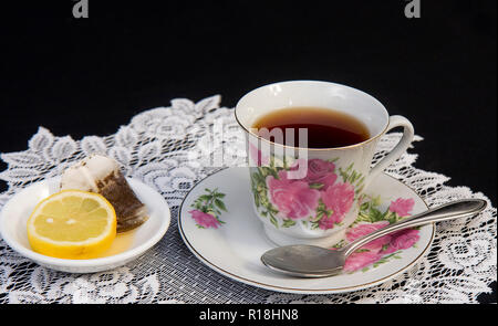 Rosa tazzina floreale con tè, fettina di limone e un sacchetto da tè mentre è seduto sul pizzo bianco centrino su uno sfondo nero. Concetto di immagine. Foto Stock