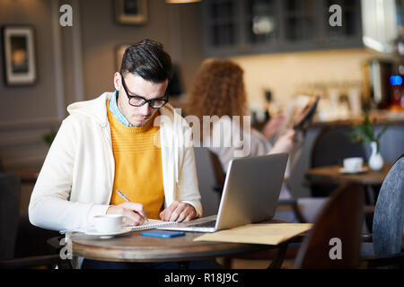 Creative giovane uomo in occhiali rendendo note in notepad stando seduti al bar nella parte anteriore del computer portatile Foto Stock
