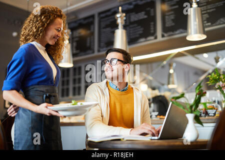 Giovane cameriera portando la fine di uno dei clienti seduti a tavola e networking Foto Stock