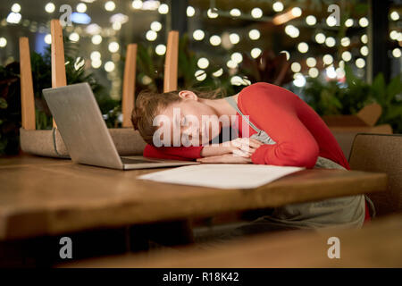 Stanco giovane donna in grembiule di dormire sulla tavola in cafe con laptop e carte nelle vicinanze Foto Stock