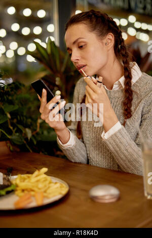 Bella ragazza casual applicando Lip Gloss dopo aver mangiato guardando nella schermata dello smartphone in cafe Foto Stock