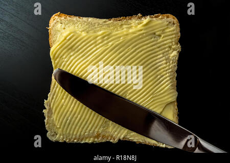 Il burro con belle macchie e un coltello su un pezzo di pane di close-up su sfondo scuro Foto Stock