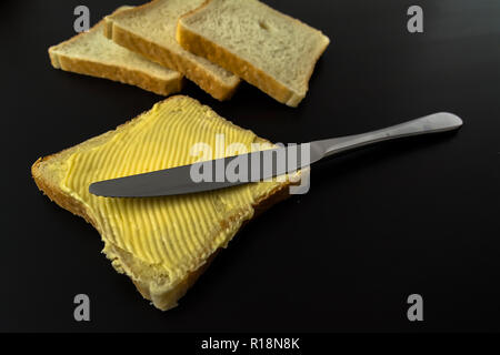 Il burro con belle macchie e un coltello su un pezzo di pane di close-up su sfondo scuro Foto Stock