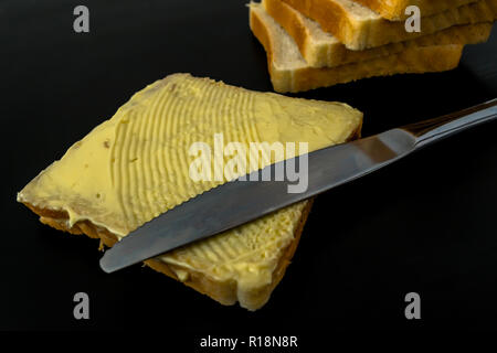 Il burro con belle macchie e un coltello su un pezzo di pane di close-up su sfondo scuro Foto Stock