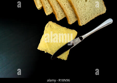 Il burro con belle macchie e un coltello su un pezzo di pane di close-up su sfondo scuro Foto Stock