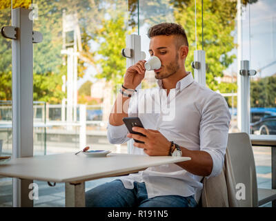 Chiusura del bel giovane di bere il caffè presso il negozio mentre guarda il Telefono Cellulare Foto Stock