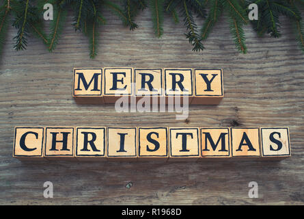 Cubi di legno con lettere formano il testo inglese Buon Natale. Essi sono giacenti su un sfondo di legno. Rami di abete di incorniciare l'immagine. Foto Stock