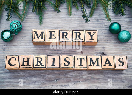 Cubi di legno con lettere formano il testo inglese Buon Natale. Essi sono giacenti su un sfondo di legno. Rami di abeti e palle di Natale sono accanto ad essa. Foto Stock